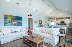 A living room with two white sofas, a coffee table, a painting of a boat, and a kitchen in the background.
