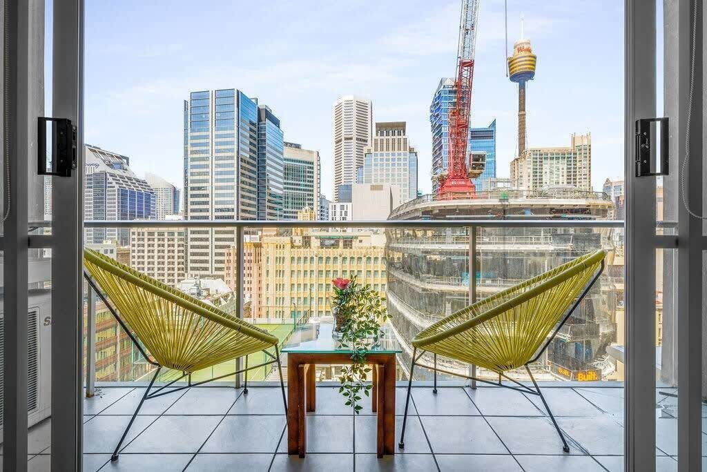 A balcony with two yellow hammocks and a table with a flower arrangement, overlooking a cityscape with modern buildings and a distinctive tower.
