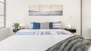 A bedroom with a bed, two bedside tables, a nightstand, a wall-mounted painting, and a window.