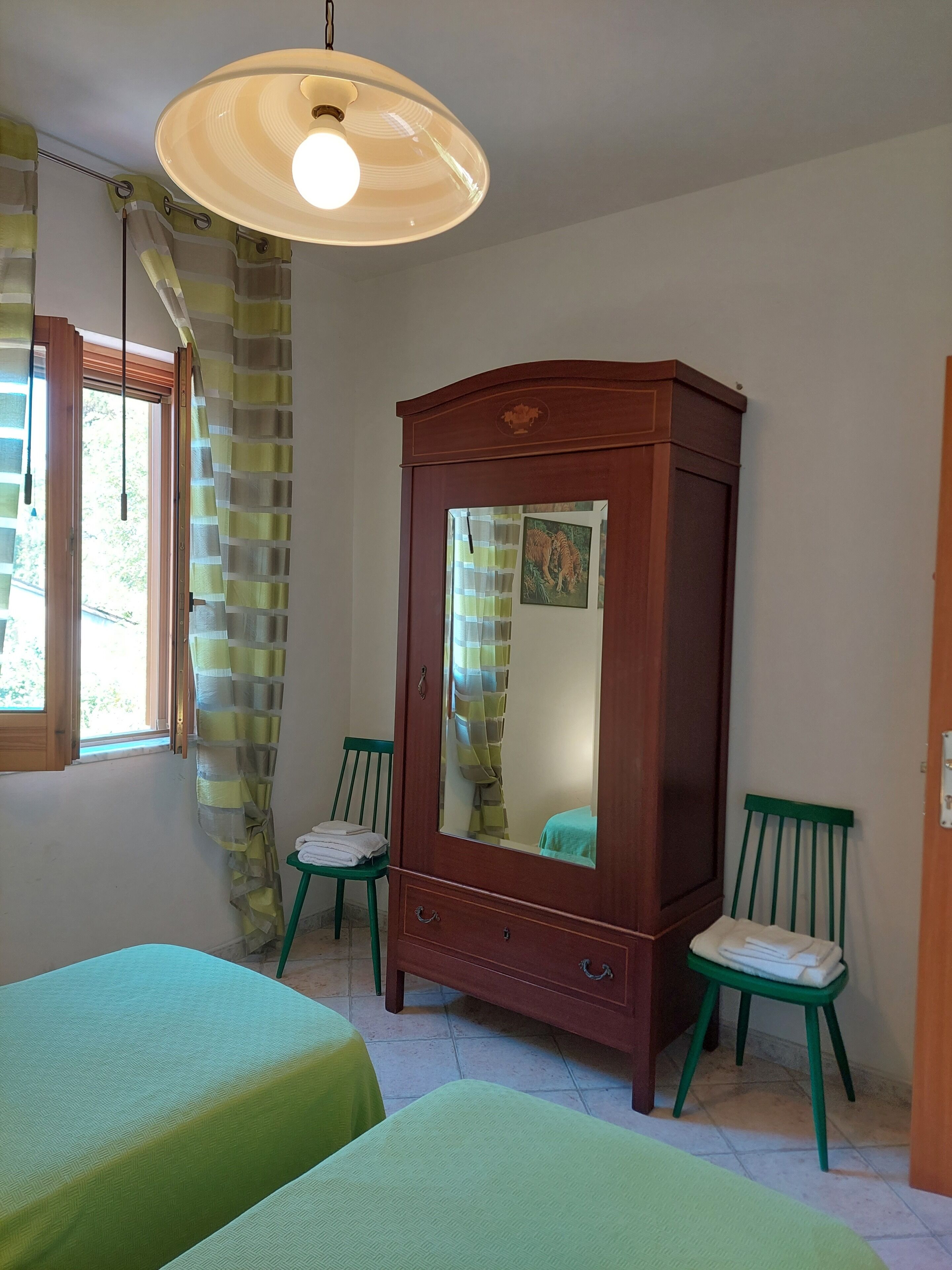 A room with two green beds, a wooden wardrobe with a mirror, and a hanging light fixture.