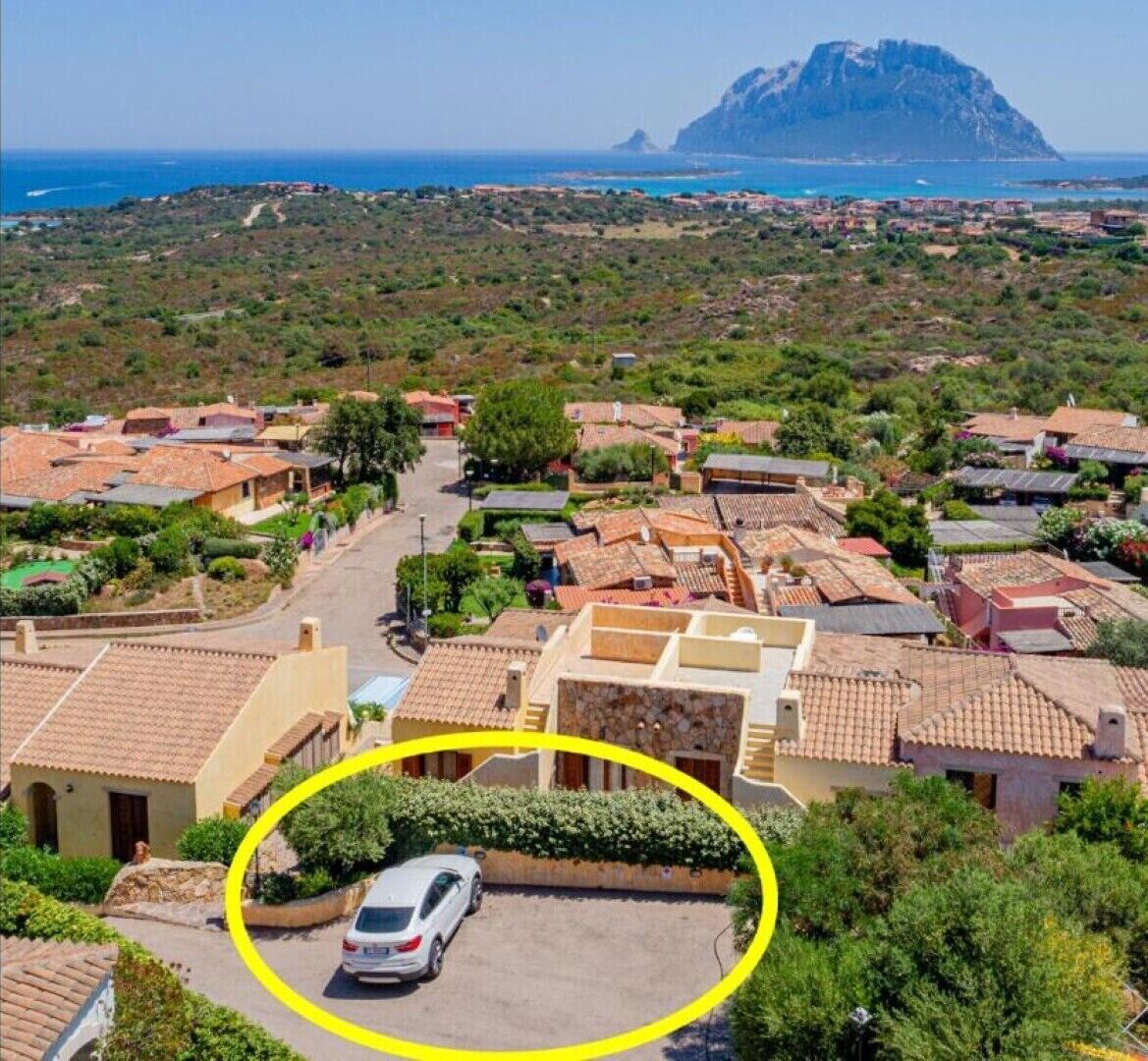 Un pueblo costero con edificios de tejados de terracota, una montaña destacada al fondo y un coche blanco estacionado en una entrada.