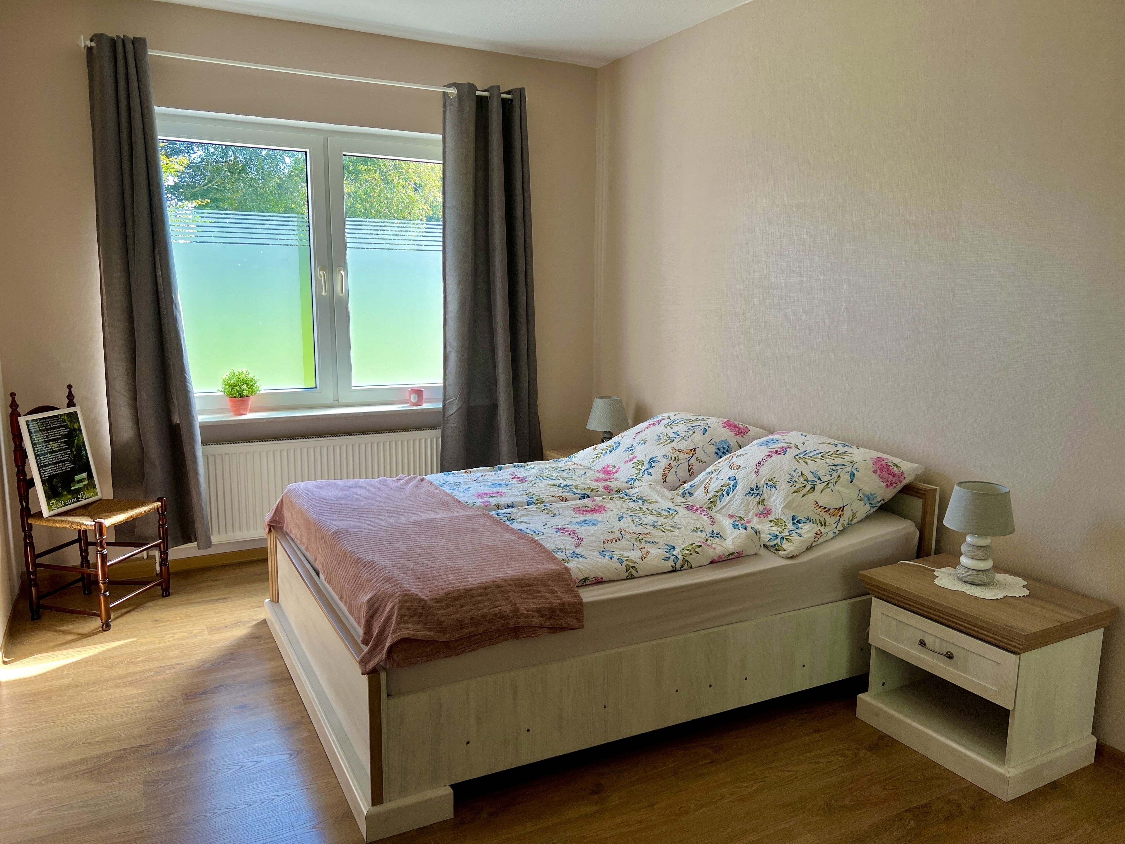 A bedroom with a bed, a nightstand, a chair, a window with curtains, and a radiator.
