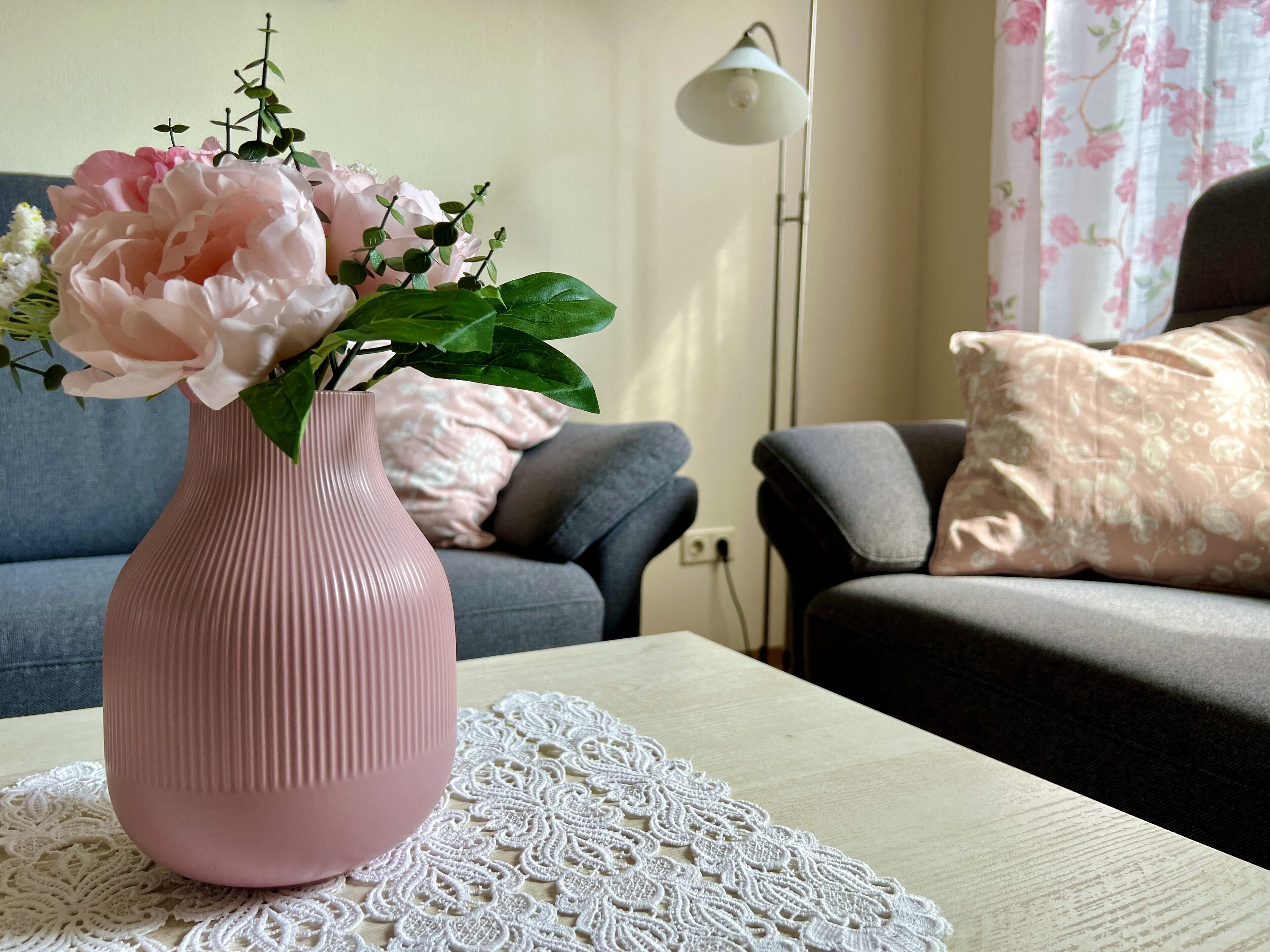 A pink vase with flowers on a table, a gray sofa, and a floor lamp in a living room.