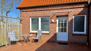 Ein Backsteinhaus mit Ziegeldach, Holzumzäunung und einer Terrasse mit zwei Stühlen und einem Tisch.