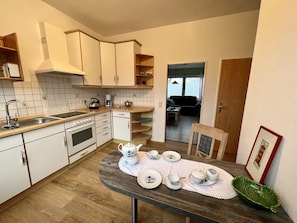 A compact kitchen with white cabinets, a sink, and a stove. There is a dining table with tea sets and a green leaf-shaped bowl.