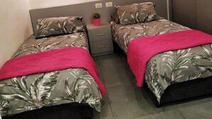 Une chambre avec deux lits, chacun recouvert d’une couvre-lit à motifs et d’une couverture rouge. Une petite plante en pot est posée sur la table de chevet, entre les deux lits.