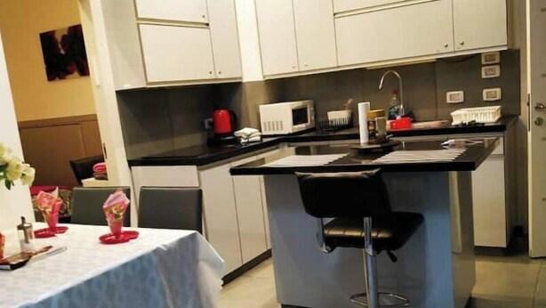 Une cuisine moderne avec des meubles blancs, un îlot central doté d’un tabouret, un four à micro-ondes et une bouilloire.