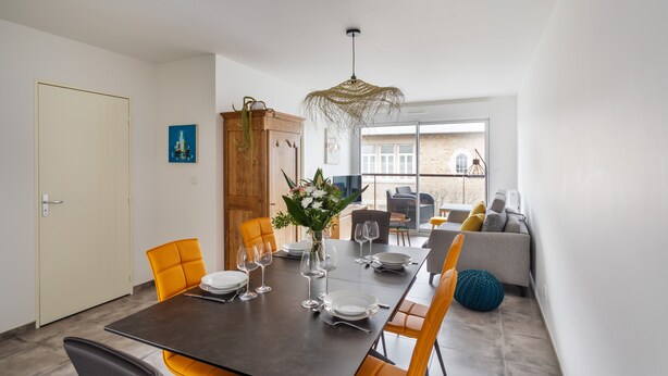 A dining area with a dark table set for six, surrounded by orange chairs. A wooden cabinet and a hanging light fixture are visible.