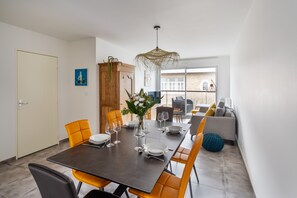 A dining area with a dark table set for six, surrounded by orange chairs. A wooden cabinet and a hanging light fixture are visible.
