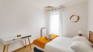 A bedroom with a bed, a desk, a radiator, a window with curtains, a wall-mounted wreath, and a lamp.