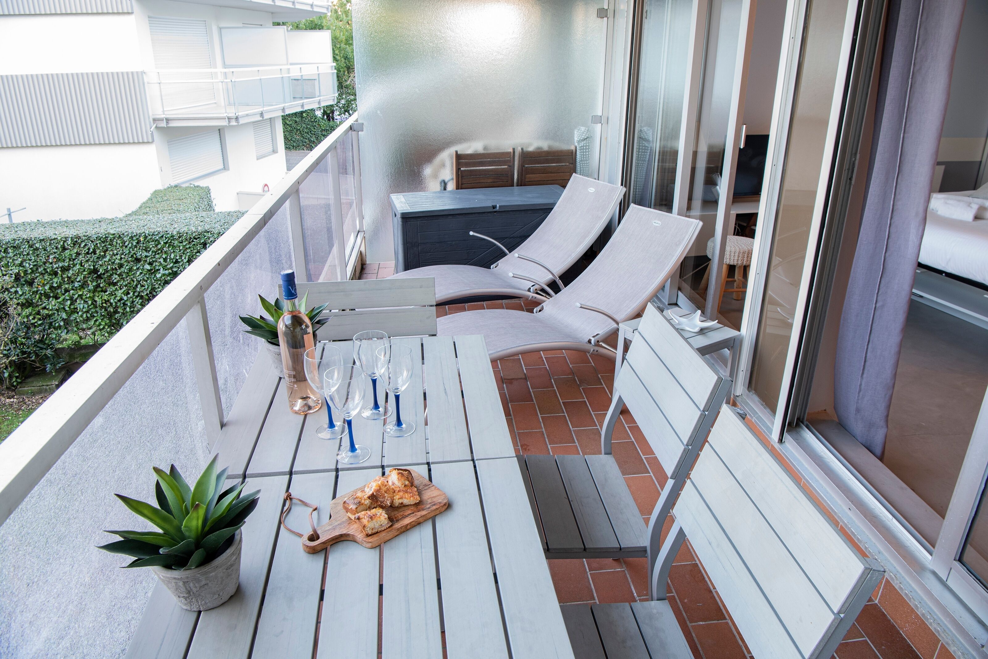A balcony with a table set for dining, two lounge chairs, a potted plant, and a sliding glass door leading to a bedroom.