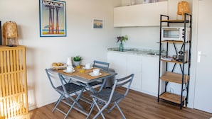 Cocina compacta con microondas, tostadora y estantes. Hay una mesa de comedor con sillas y un mueble pequeño.