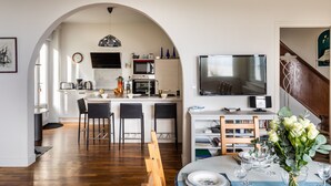 Une cuisine moderne avec un îlot central, un coin repas avec une table dressée pour deux personnes, et un téléviseur fixé au mur.