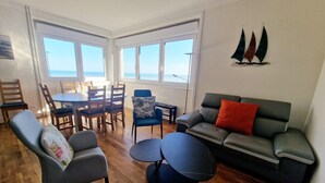 Un salon comprenant un canapé, des fauteuils, une table à manger et offrant une vue sur la mer grâce à de grandes fenêtres.