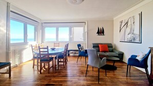 Un vaste espace de vie doté d’un plancher en bois, d’une table à manger avec des chaises, d’un canapé et offrant une vue sur la mer grâce à de grandes fenêtres.