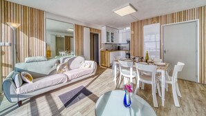 En moderne stue med sofa, spisebord og stole. Der er et køkkenområde med skabe og et køleskab.