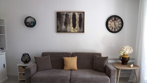 Ein Wohnzimmer mit grauem Sofa, einem an der Wand befestigten abstrakten Gemälde, einer runden Uhr und einem kleinen Beistelltisch mit Blumen in einer Vase.