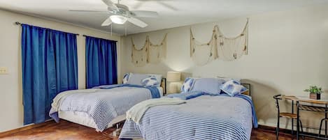 A room with two beds, a ceiling fan, a small table with a chair, and a wall-mounted light fixture.