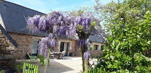 Un edificio de piedra con un porche cubierto, mesas al aire libre y enredaderas moradas con flores.