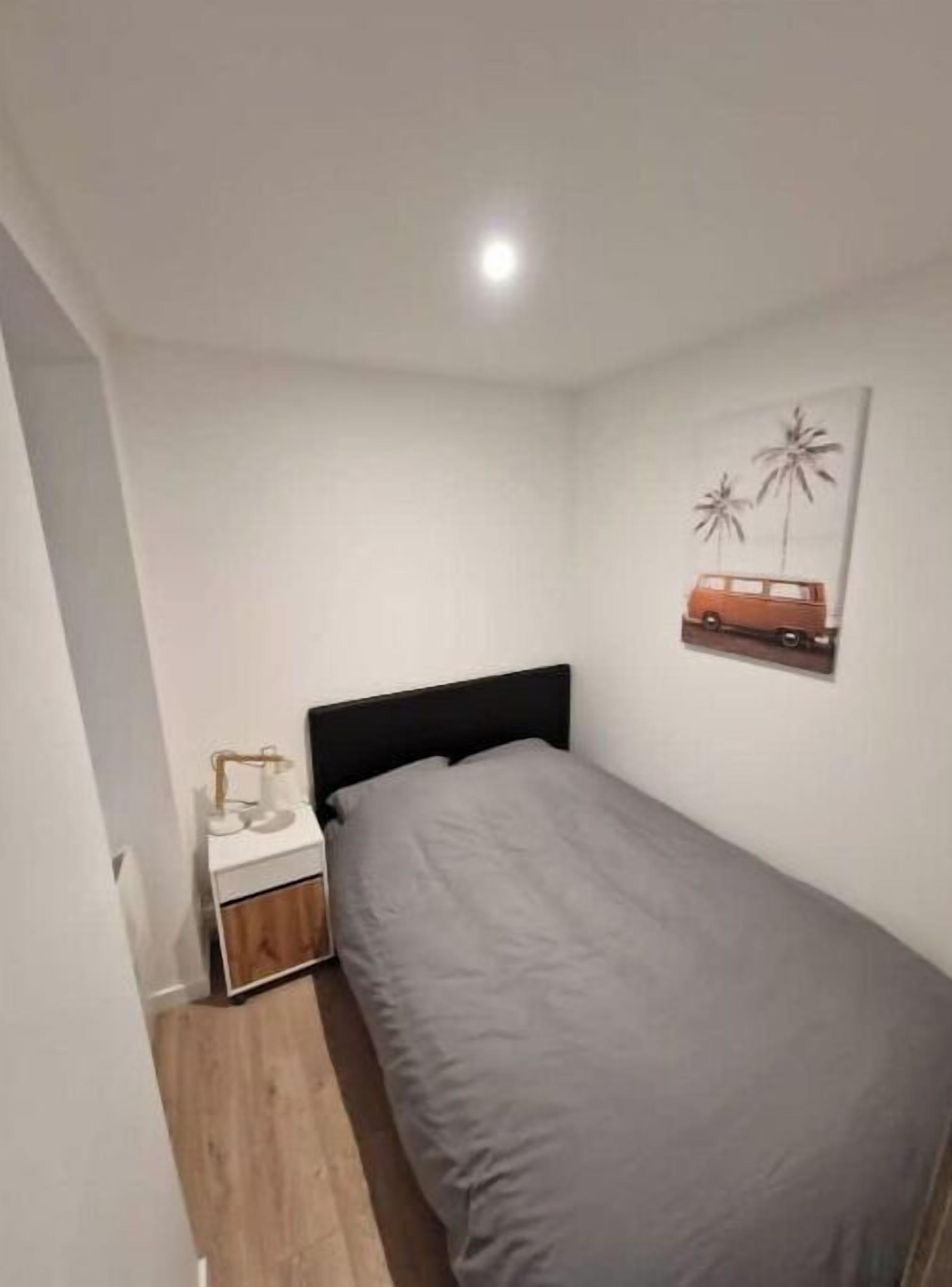 Quarto pequeno e bem organizado com uma cama de solteiro, uma mesa de cabeceira e um quadro na parede com um carro e palmeiras.