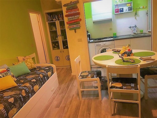 Ein kompakter Wohnbereich mit einer Couch, einem Esstisch mit Stühlen, einer Küchenzeile und einem Bücherregal.