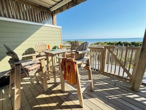 A wooden deck with outdoor furniture, including chairs and a table, overlooking a beach.