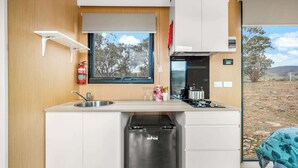 A compact kitchen with a built-in dishwasher, a window with a view of trees, and a small sink. - Cabernet Tiny House by Tiny Away (Windeyer)