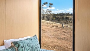 A room with a large window, a bed with a green patterned pillow, and a view of a dry landscape with sparse trees and hills. - Cabernet Tiny House by Tiny Away (Windeyer)