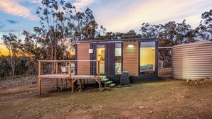 A small, modern cabin with a wooden deck, a large water tank, and a view of the surrounding trees. - Cabernet Tiny House by Tiny Away (Windeyer)