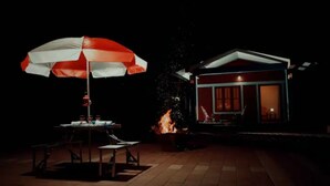 A patio with a table and chairs under an umbrella, a small fire pit, and a house with a lit interior. - Entire Villa at the Heart of the Wayanad Forest. (Thirunelly)