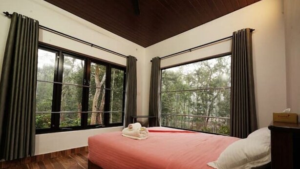 A bedroom with a bed, two windows, and a view of trees. - Entire Villa at the Heart of the Wayanad Forest. (Thirunelly)