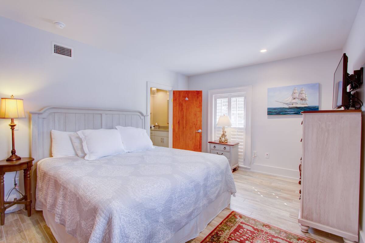 A bedroom with a large bed, a nightstand, a dresser, a door, a painting, and a window.