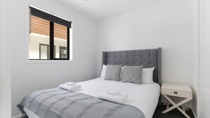Een moderne slaapkamer met een grijs beklede hoofdeinde, een net opgemaakt bed met wit en grijs beddengoed, een klein nachtkastje en een raam met zonwering.