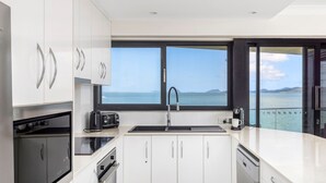 Une cuisine moderne avec des meubles blancs, un plan de travail noir et une vue sur la mer grâce à une grande fenêtre.