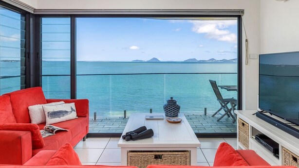 Un salon moderne avec un canapé rouge, une table basse et un téléviseur. La pièce est dotée de grandes fenêtres qui offrent une vue sur la mer et les montagnes lointaines.