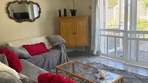 A living room with two grey sofas, red and grey cushions, a wooden coffee table, a ceiling fan, a wooden cabinet, and a large window with a view of greenery. - Sizzling Hot Summer so stay by the Beach (New Smyrna Beach)