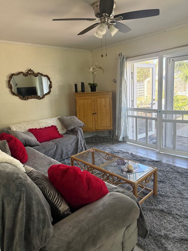 A living room with two grey sofas, red and grey cushions, a wooden coffee table, a ceiling fan, a wooden cabinet, and a large window with a view of greenery.