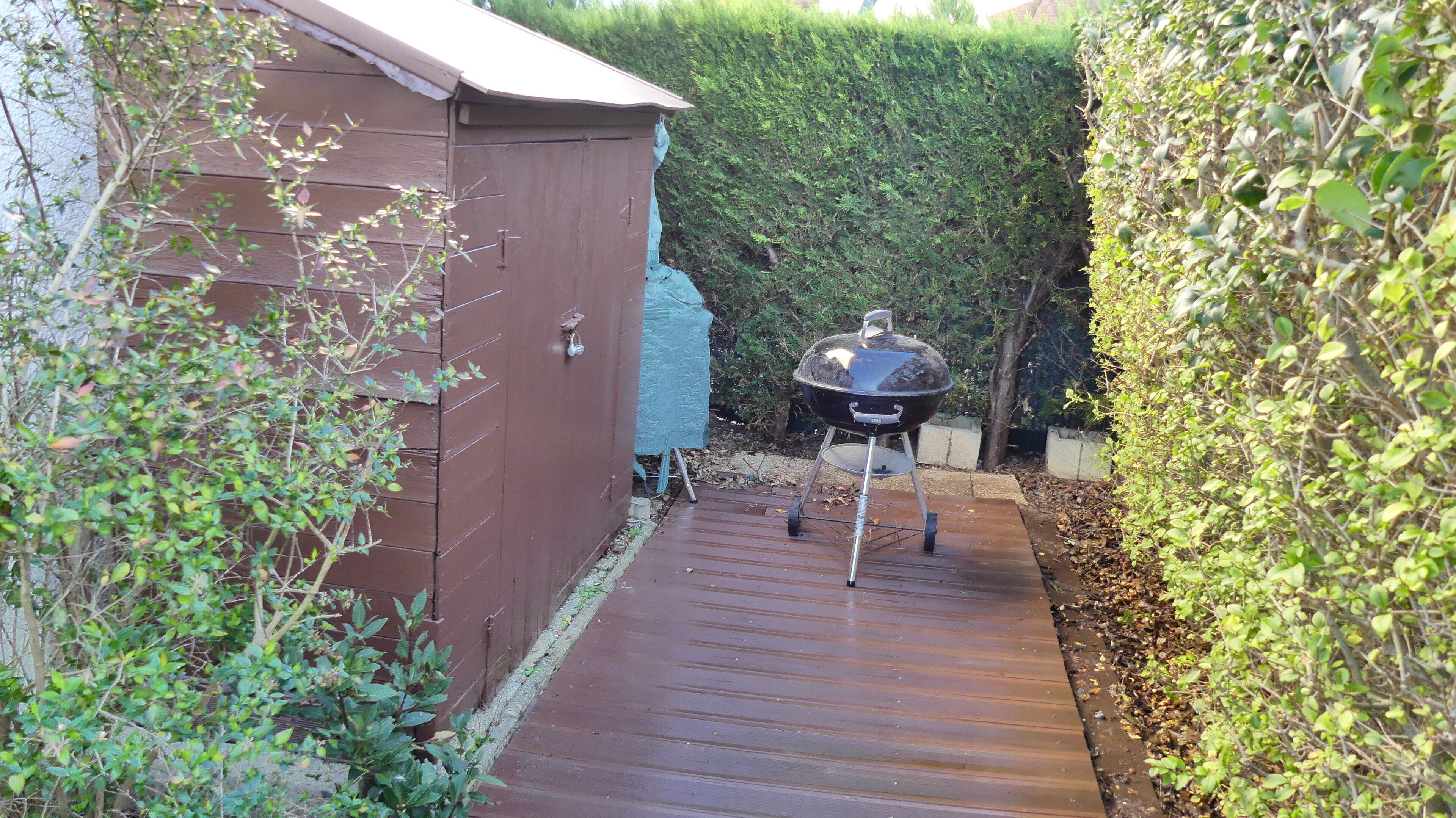 A wooden deck leading to a barbecue grill, a shed, and a hedge.
