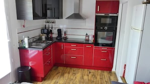 A compact kitchen with red cabinets, a stainless steel sink, and a built-in oven. - Charming sunny apartment | Les Manoirs Normands (Courseulles-sur-Mer)