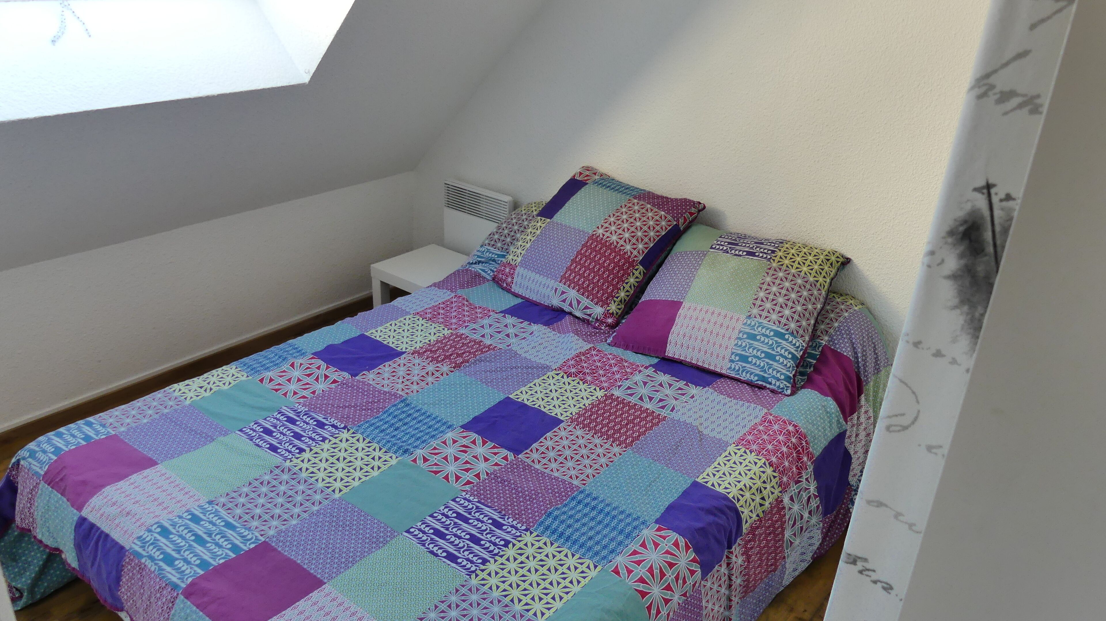 Quarto com colcha de retalhos colorida, mesa de cabeceira branca e janela com vista para o céu.