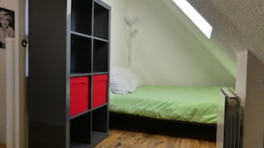 A small, single-bed room with a sloped ceiling, a radiator, and a bookshelf with red and black compartments.