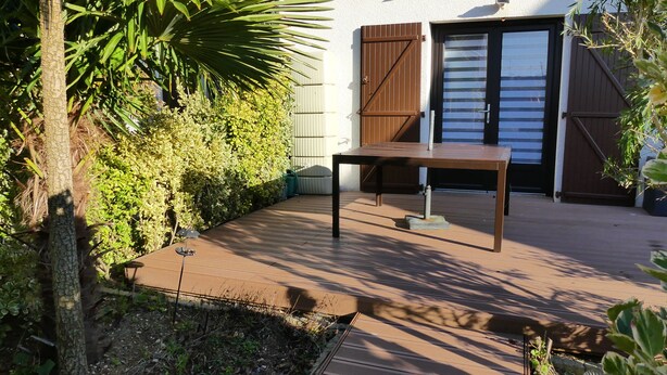 A patio with a table and chairs, surrounded by greenery and a building with a brown door. - Charming sunny apartment | Les Manoirs Normands (Courseulles-sur-Mer)