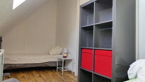 A small, compact room with a sloped ceiling, a bed, a small table with a lamp, and a red storage unit. - Charming sunny apartment | Les Manoirs Normands (Courseulles-sur-Mer)