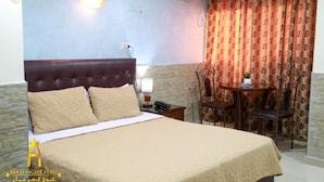 Quarto de hotel com cama, mesa de cabeceira com luminária, uma cadeira e uma pequena mesa com uma planta.