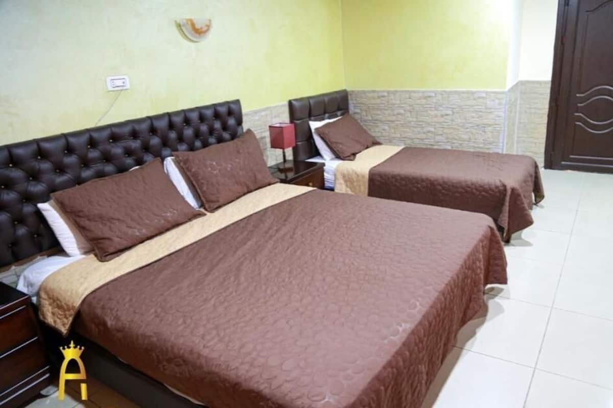 Quarto de hotel com duas camas, uma mesa de cabeceira, uma luminária e uma porta.