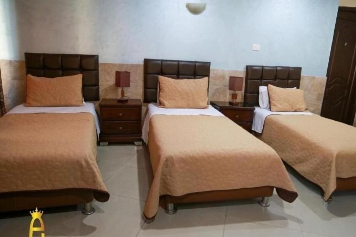 A hotel room with three beds, each with a brown headboard and matching bedding. There are bedside tables with lamps and a wall-mounted light fixture.