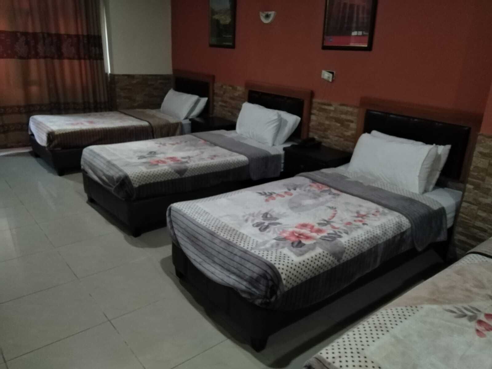 A hotel room with three single beds, each with a floral bedspread and a pillow. The room has a tiled floor and a brick accent wall.