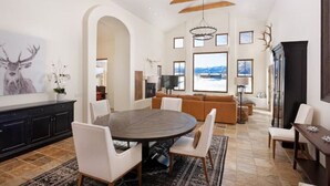 A spacious living area with a round dining table, white chairs, a brown sofa, and a large window offering a view of the mountains.