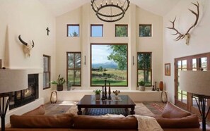 Relax in a spacious living room with a high ceiling and scenic views, featuring a comfortable seating area, fireplace, and decorative elements.