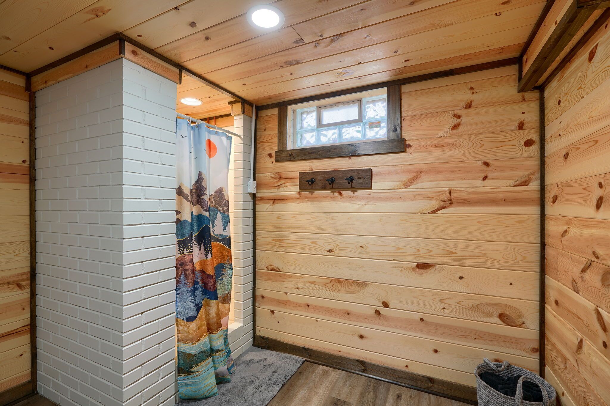 A wooden cabin interior with a shower curtain featuring a mountain and forest design, a small window, and a wooden bench.